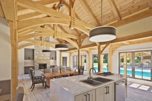 An interior shot from the BigSkyHaven floor plan, the latest addition to the Woodhouse catalog. The open space combines a kitchen, dining room, and living room under high, vaulted timber wood frame ceilings. The room is lit by three hanging circular lamps as well as large, floor to ceiling windows that look out onto a pool.