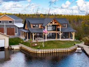 A lakeside timber frame home with a wraparound covered porch, large windows, and dockside landscaping on a calm waterfront.