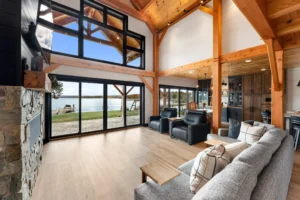 Spacious timber frame great room with floor-to-ceiling windows, stone fireplace, exposed beams, and panoramic lake views.