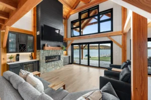Timber frame great room with exposed wood beams, stone fireplace, and full-height windows overlooking a lakefront patio and fall shoreline.
