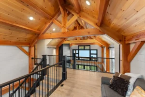 Open loft with timber frame trusses and black railing overlooking the great room and expansive lake views through floor-to-ceiling windows.