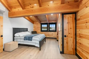 Lake-view bedroom with tongue-and-groove wood walls, exposed ceiling beams, black-framed windows, and a simple modern bed.