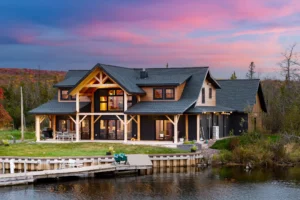 Timber frame waterfront home at sunset, featuring glowing interior lighting, covered patios, and a wooded backdrop with fall colors.