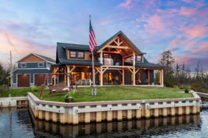 Front view of a modern timber frame home on the water, highlighting exposed wood beams, expansive windows, and a grassy shoreline.