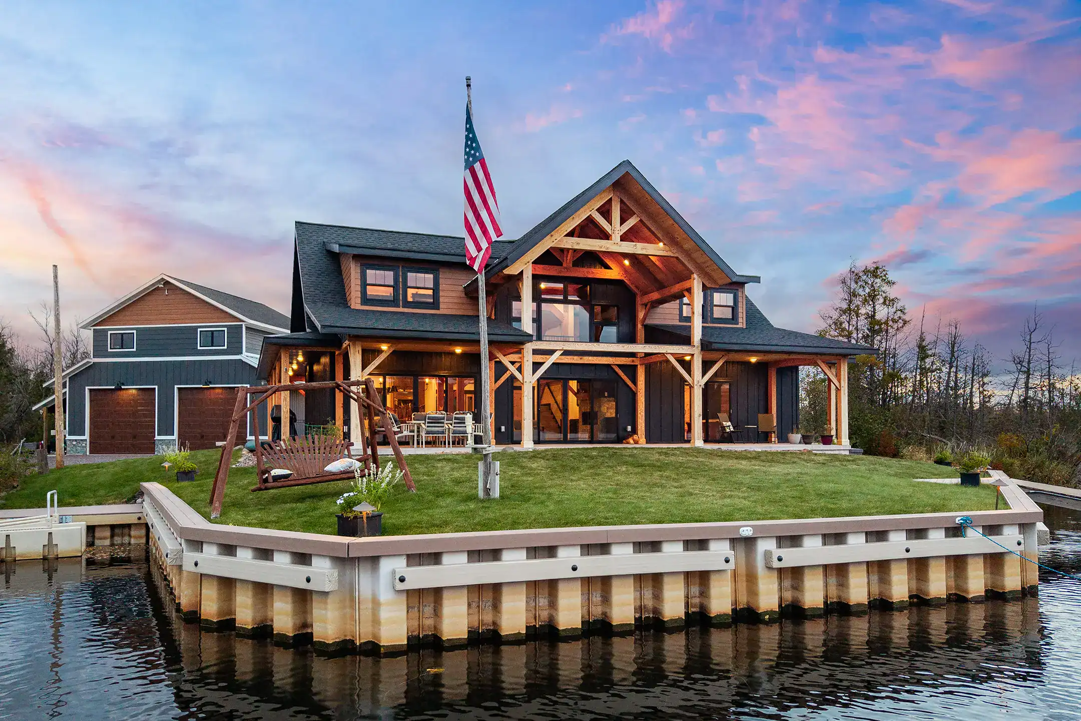 Front view of a modern timber frame home on the water, highlighting exposed wood beams, expansive windows, and a grassy shoreline.