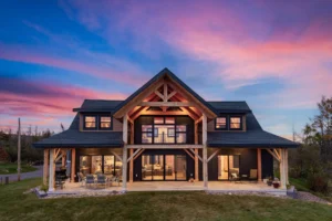 Centered view of a timber frame home at dusk, showcasing a gabled entry, illuminated interior spaces, and a stone-edged patio.