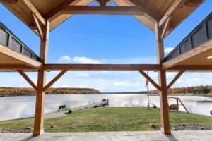 View from beneath a timber frame porch looking out toward a calm Michigan lake with a dock, fall foliage, and clear blue sky.