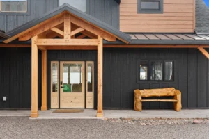 Front entry of a timber frame home featuring natural wood posts, gabled entry roof, black vertical siding, and a handcrafted log bench.