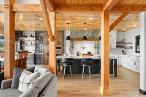 Open-concept timber frame kitchen and dining area with exposed wood beams, stone range hood, large island, wood cabinetry, and warm natural finishes.