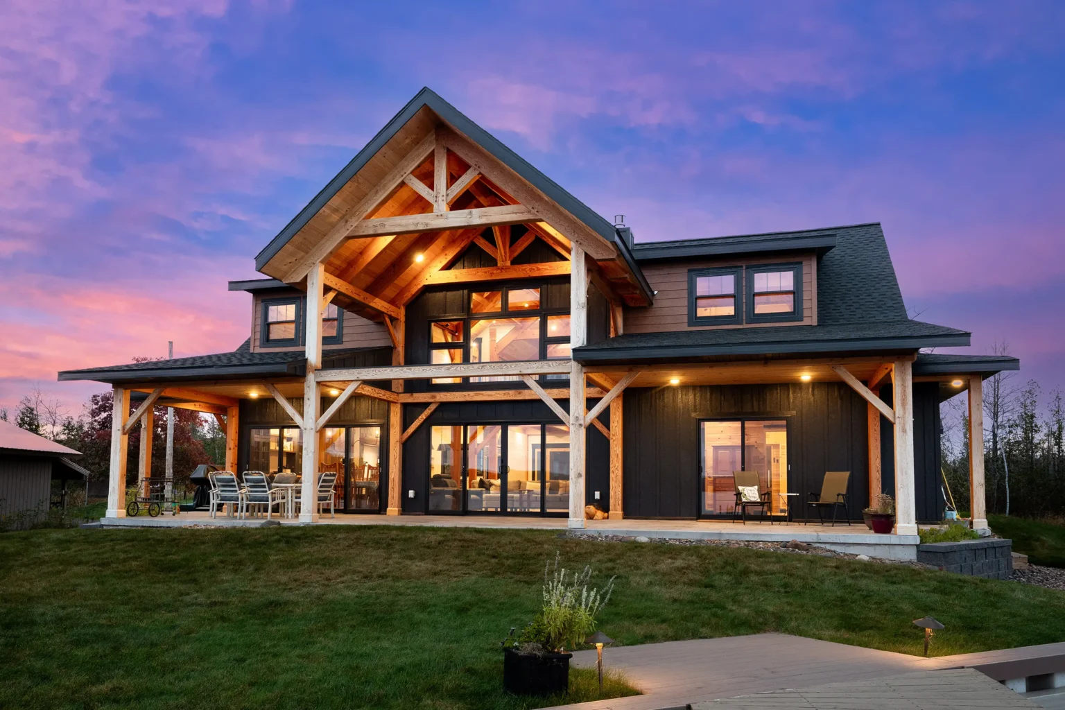 Exterior of a timber frame home at sunset with large gabled porch, warm interior lighting, and covered patio facing a grassy yard.