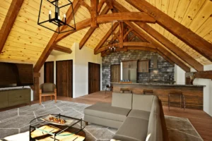 Interior of the BronsonV2’s open-concept great room, featuring vaulted timber trusses, a light wood plank ceiling, and a stone accent wall behind the kitchen. A sectional sofa faces a mounted TV, with a round coffee table centered on a geometric rug. The kitchen includes a stone countertop bar, wood cabinetry, and modern appliances. Natural light enhances the warm textures of the exposed beams and hardwood floors.