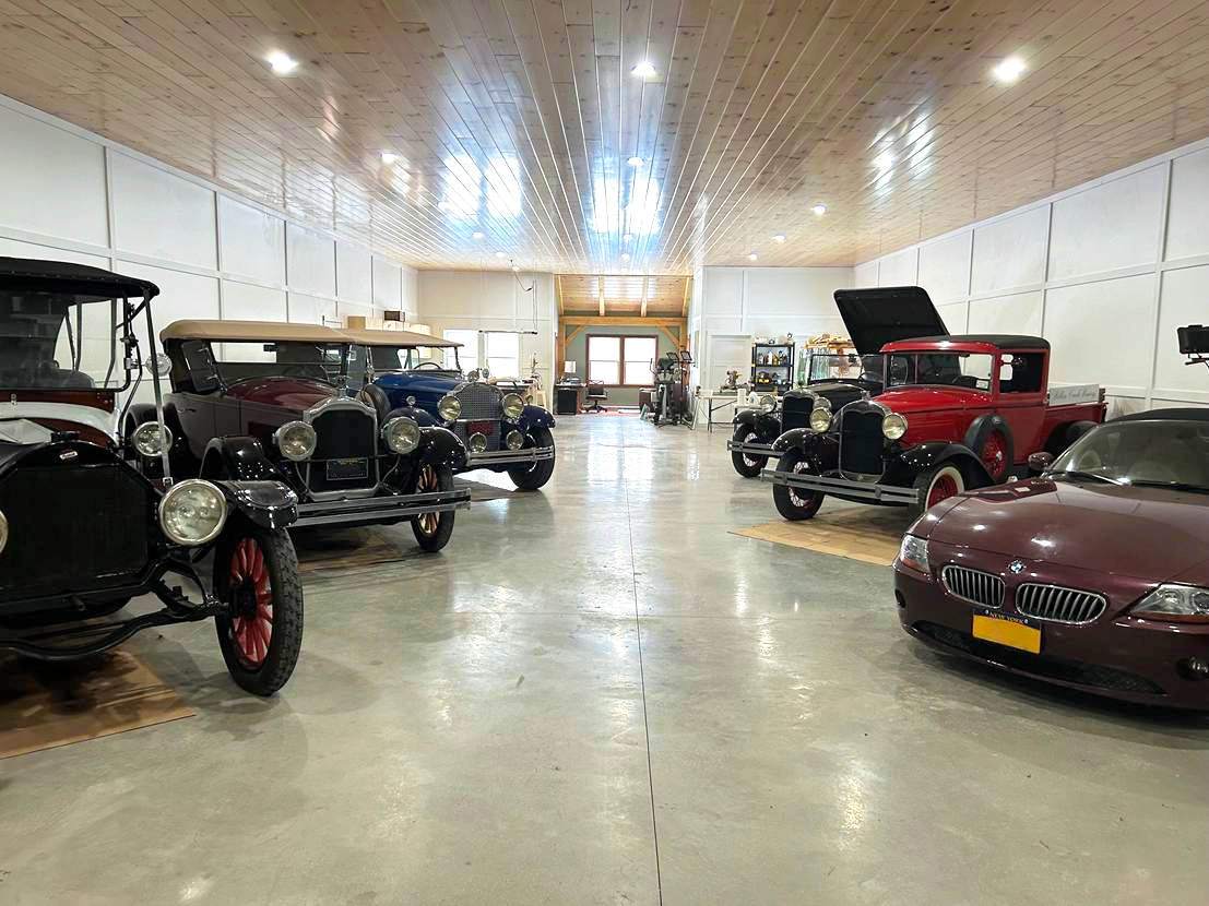 Interior of a clean, well-lit car barn with polished concrete floors and a wood plank ceiling, displaying several restored vintage automobiles from the early 20th century parked in rows, including classic sedans and coupes in black, blue, and red.