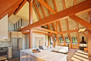 Interior view of the AspenValley A-frame home showing timber frame structure, kitchen island, loft above, skylights, and wood stove within a bright open living space.