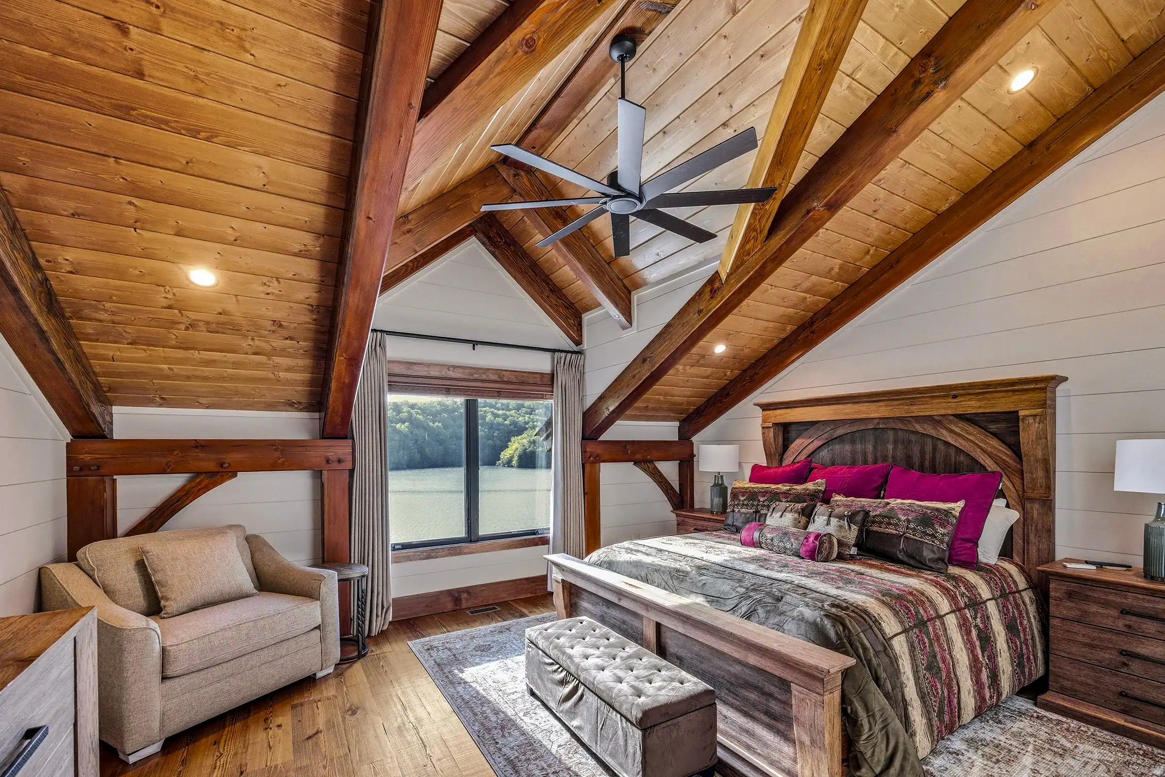 Luxury timber frame bedroom with exposed wood beams, vaulted ceilings, warm natural finishes, and expansive lake views through large windows.