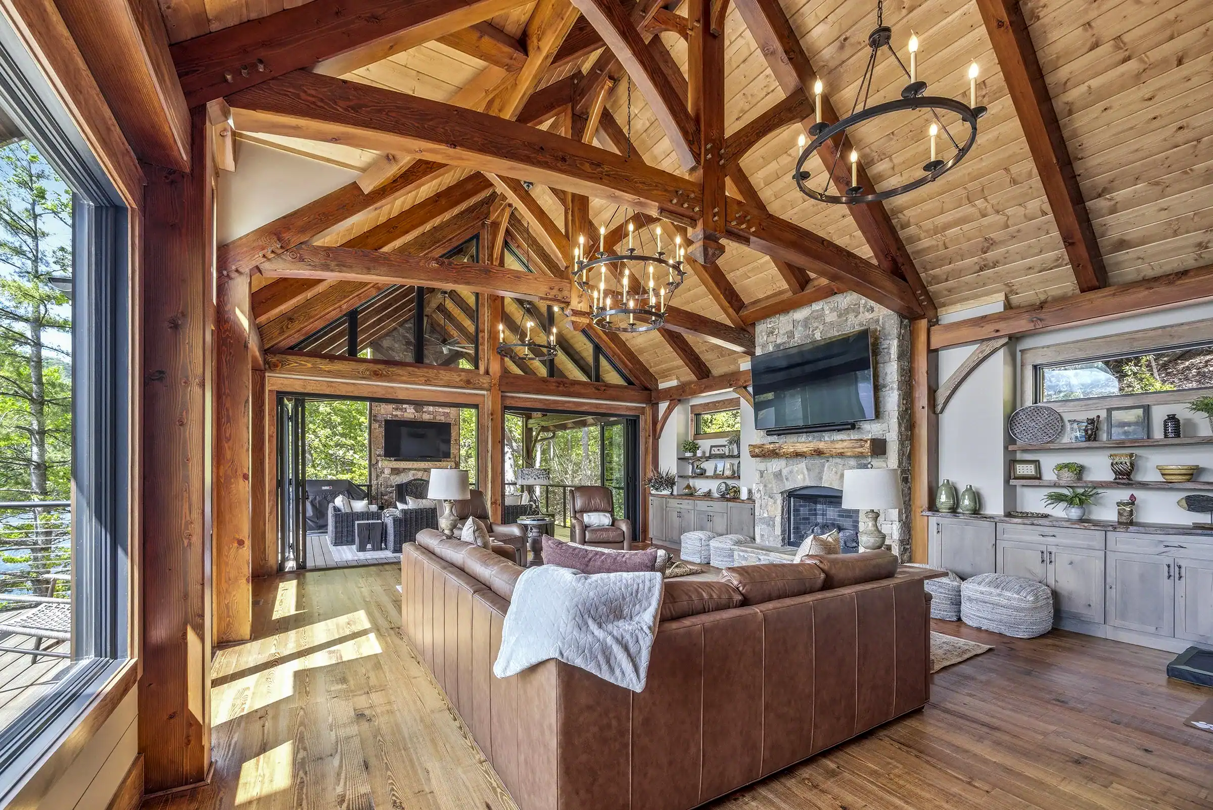 Luxury timber frame great room interior with exposed wood trusses, vaulted ceilings, natural stone fireplace, and expansive windows overlooking the lake.