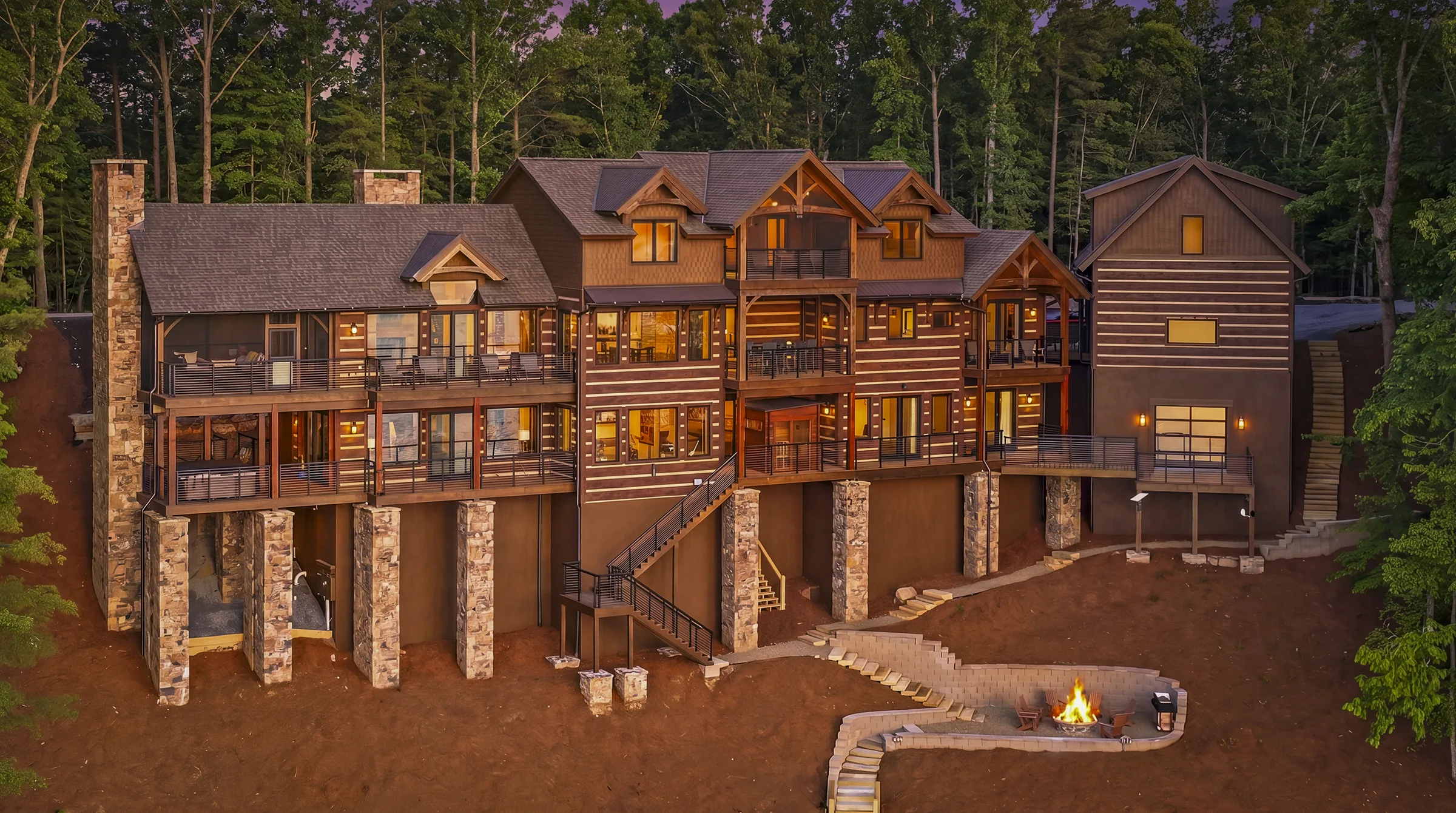 Luxury timber frame lake home aerial closeup at twilight with exposed wood framing, illuminated outdoor living spaces, expansive windows, and panoramic waterfront views.