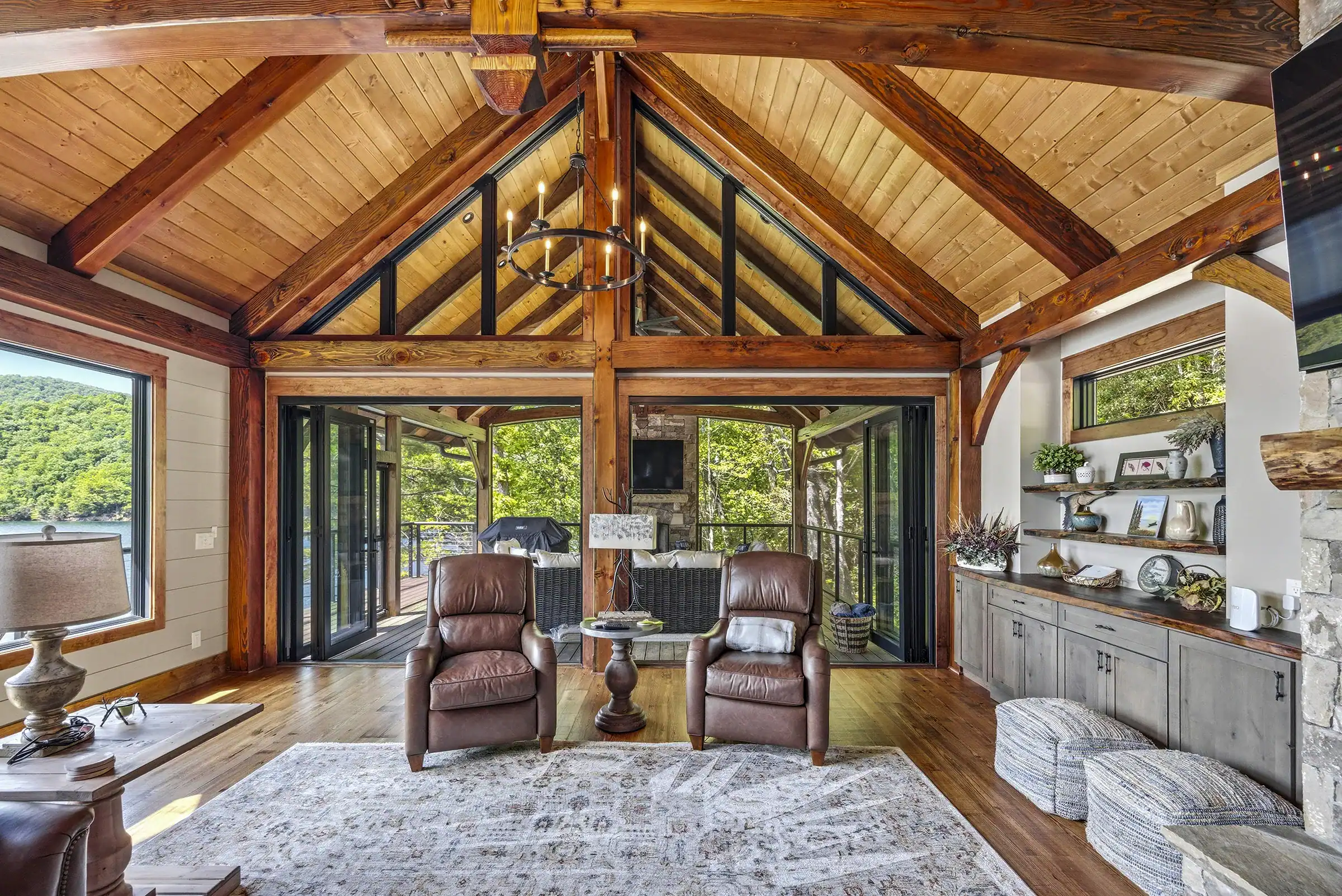 Luxury timber frame great room with vaulted ceilings, exposed wood trusses, floor-to-ceiling windows, and panoramic lake views.