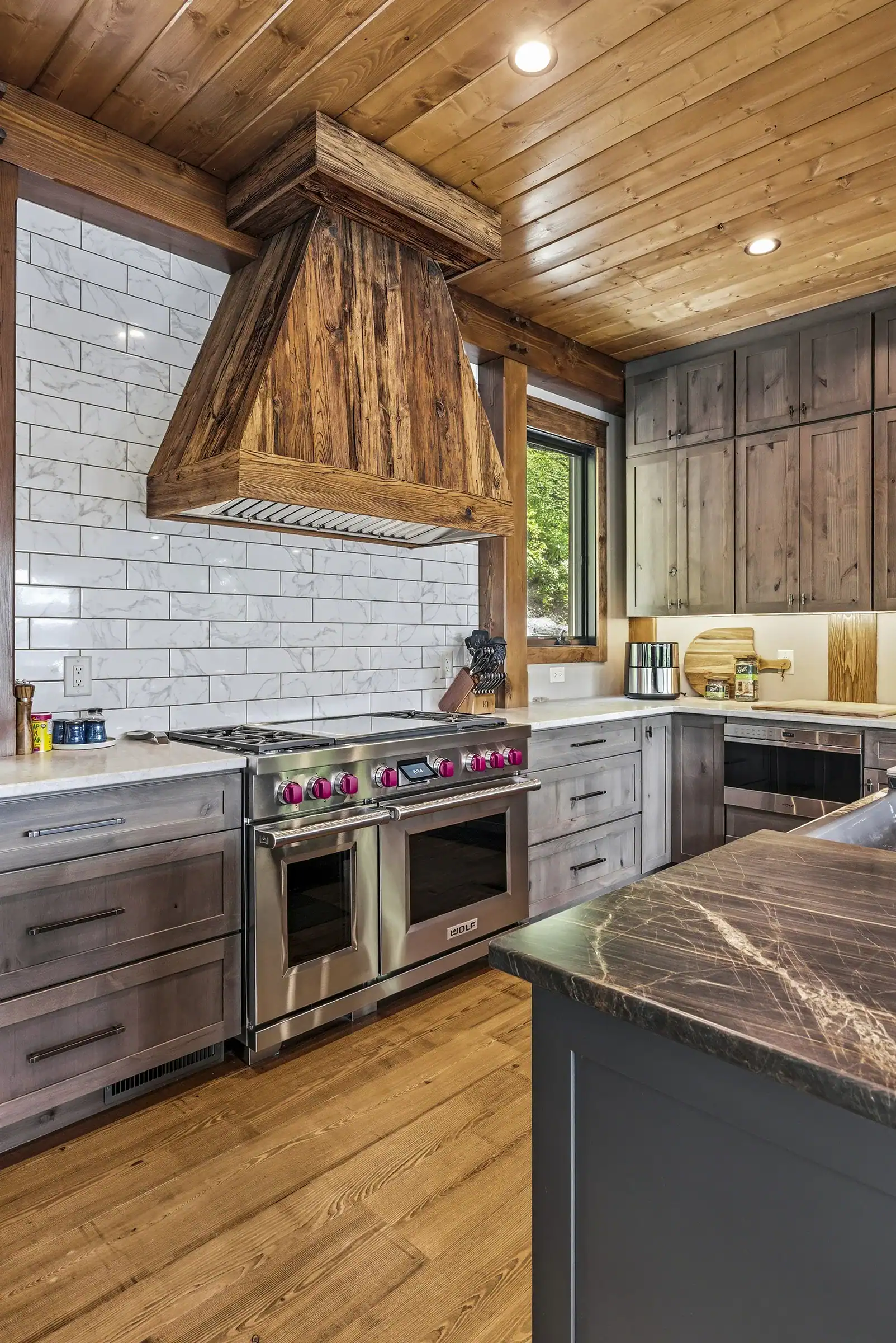 Luxury timber frame kitchen with exposed wood beams, custom wood range hood, natural stone backsplash, and large center island with seating.
