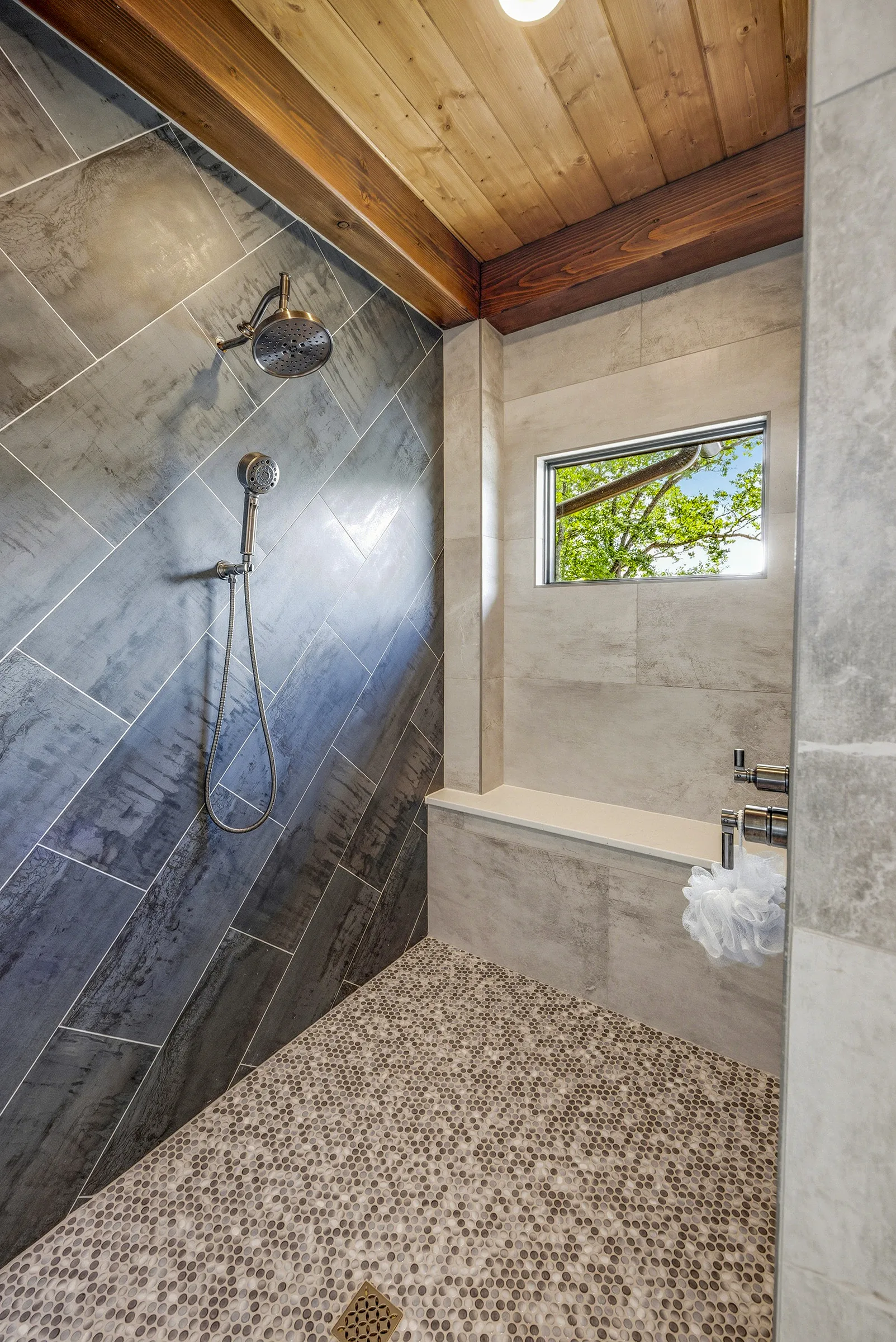 Walk-in tile shower with pebble stone flooring, large-format gray tile walls, and warm timber frame ceiling accents in a luxury mountain home bathroom.