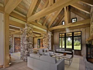 Open timber frame great room with exposed wood beams, vaulted ceiling, stone support pillars, fireplace, and seating area filled with natural light.