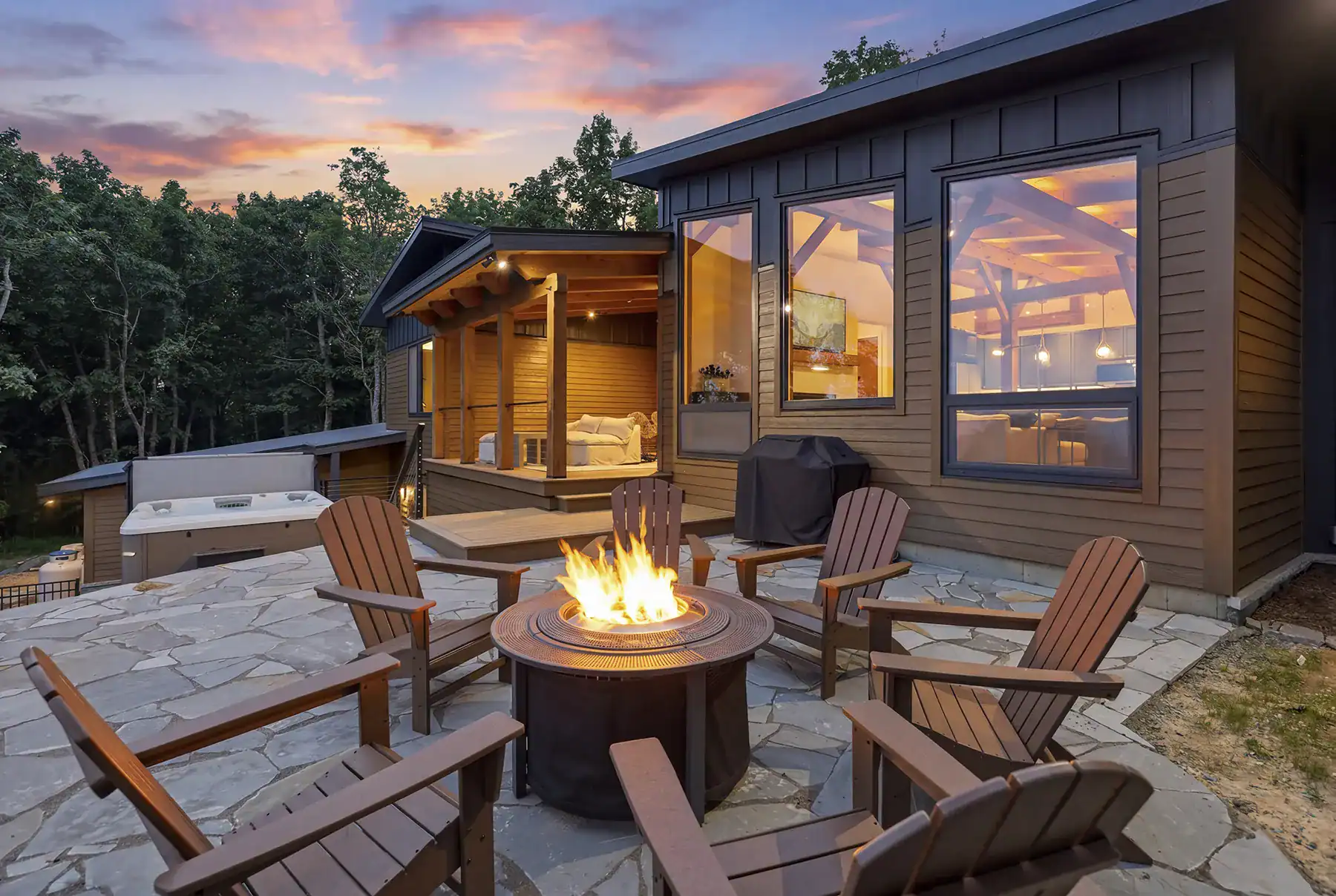 Stone patio with a fire pit, Adirondack chairs, and outdoor living area beside a custom contemporary timber frame home at twilight on Snakeden Mountain.