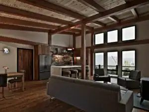 Open-concept great room in the RiverRock V2 timber frame home with exposed wood beams, floor-to-ceiling windows, stone accent wall, and modern kitchen.