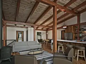 Open-concept living room and kitchen in the RiverRock V2 timber frame home featuring exposed timber beams, wood stove, large island, and modern finishes.
