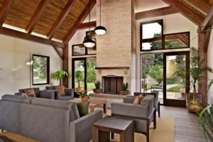 Vaulted great room in the Palmetto Sanctuary with exposed timber framing, a floor-to-ceiling brick fireplace, large windows, and doors opening to the outdoor living area.