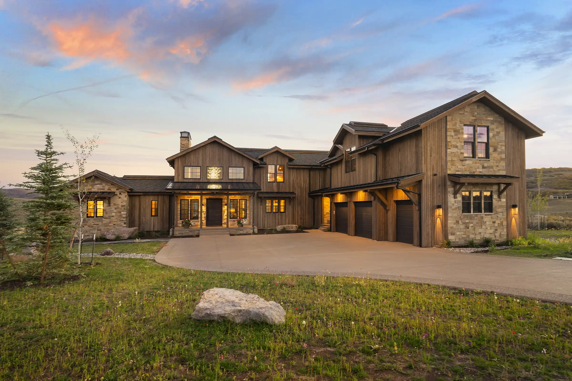 Custom timber frame home at sunset featuring natural wood siding, stone accents, a three-car garage, and a landscaped driveway beneath a colorful evening sky.