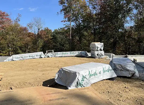 Woodhouse timber frame building materials wrapped and staged on a cleared construction site surrounded by trees.