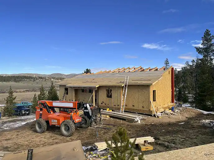 Custom timber frame home under construction on a sloping rural property, with an orange telehandler, building materials, and wooded terrain.