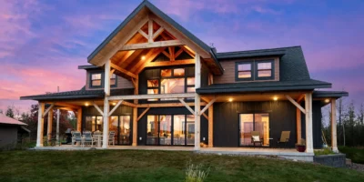 Exterior of a timber frame home at sunset with large gabled porch, warm interior lighting, and covered patio facing a grassy yard.